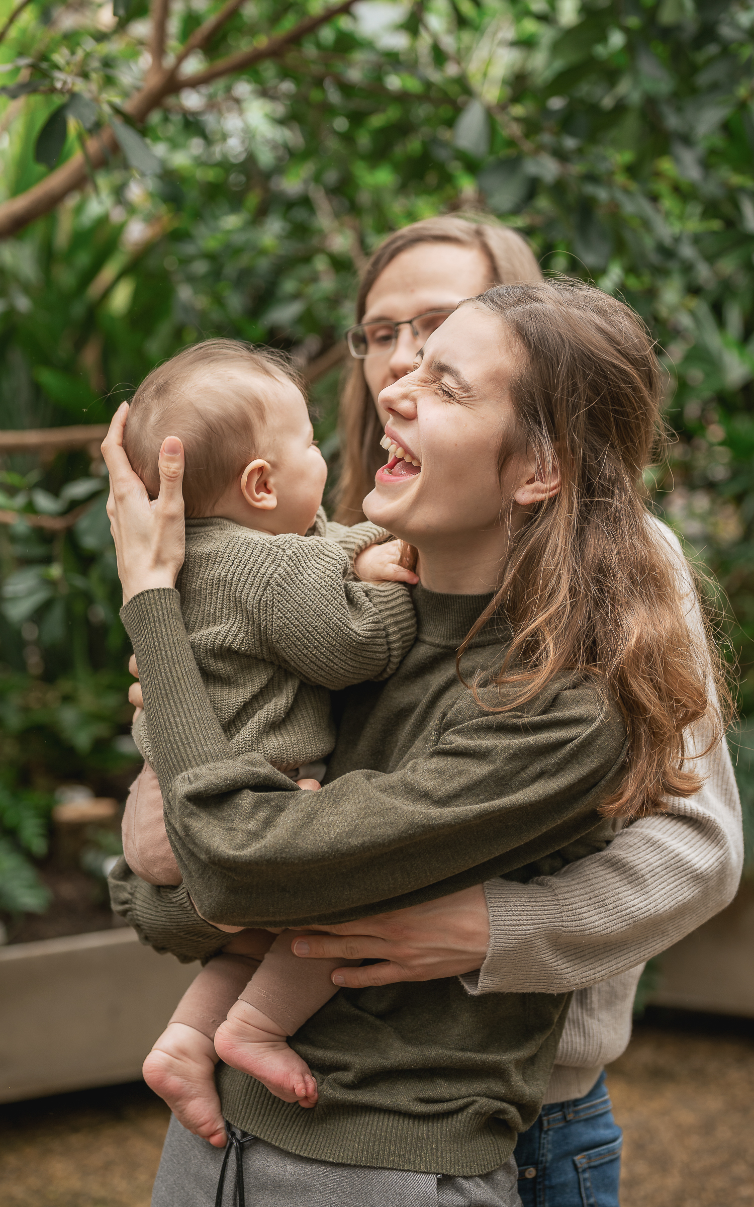 Herzlich lachende junge Mutter wird von Ehemann umarmt und hat das Baby auf dem Arm