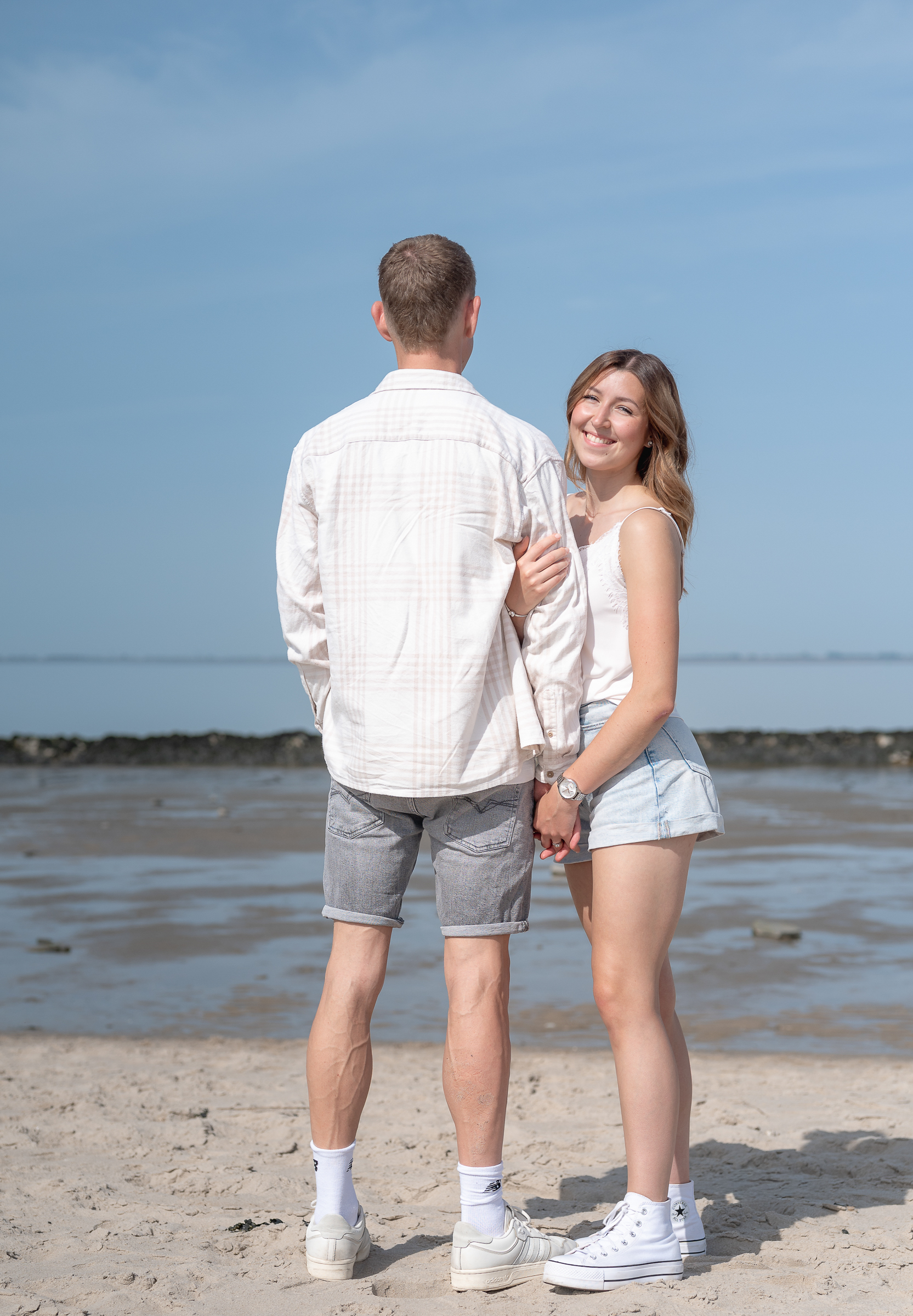Ein Paar steht am Strand, er schaut auf das Wasser, sie in die Kamera